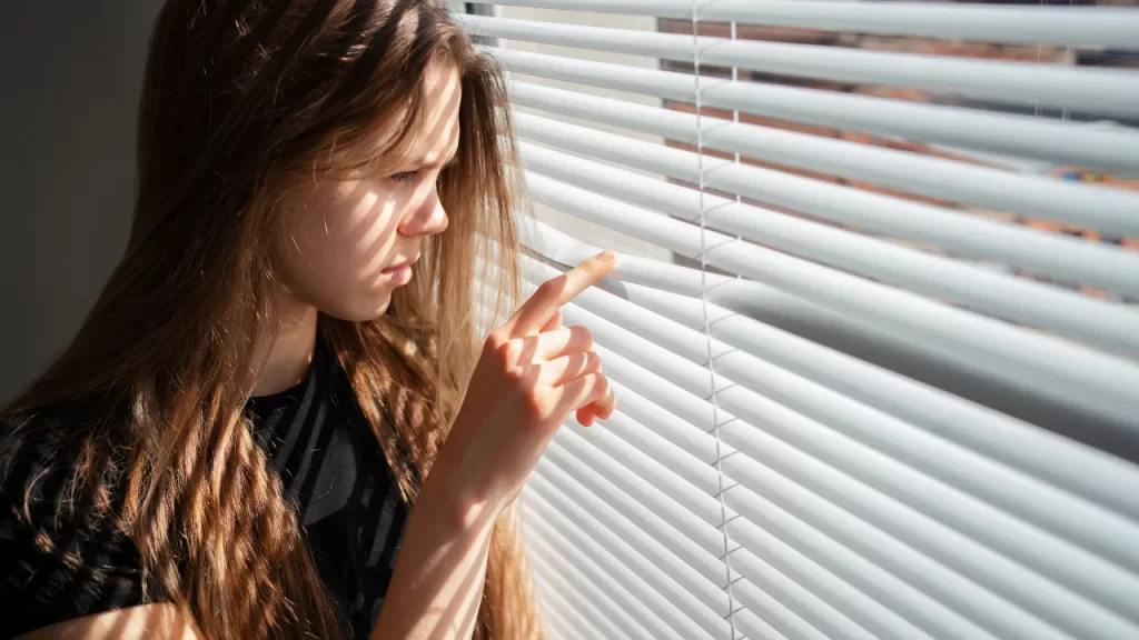 Mulher observa através de persiana horizontal com luz do sol incidindo, ambiente interno iluminado parcialmente.
