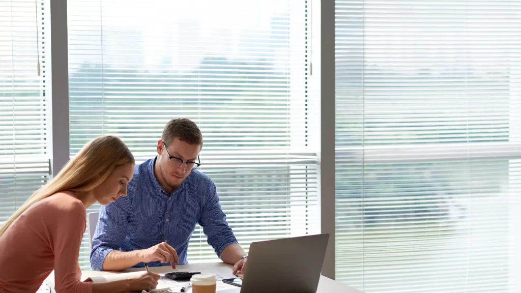 Dois profissionais trabalhando em um escritório moderno, com laptop e documentos sobre a mesa, e ao fundo uma persiana para escritório branca controlando a entrada de luz natural.