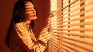 Mulher iluminada pela luz do sol filtrada por persianas, simbolizando o controle de luminosidade oferecido pelas persianas blackout motorizadas.